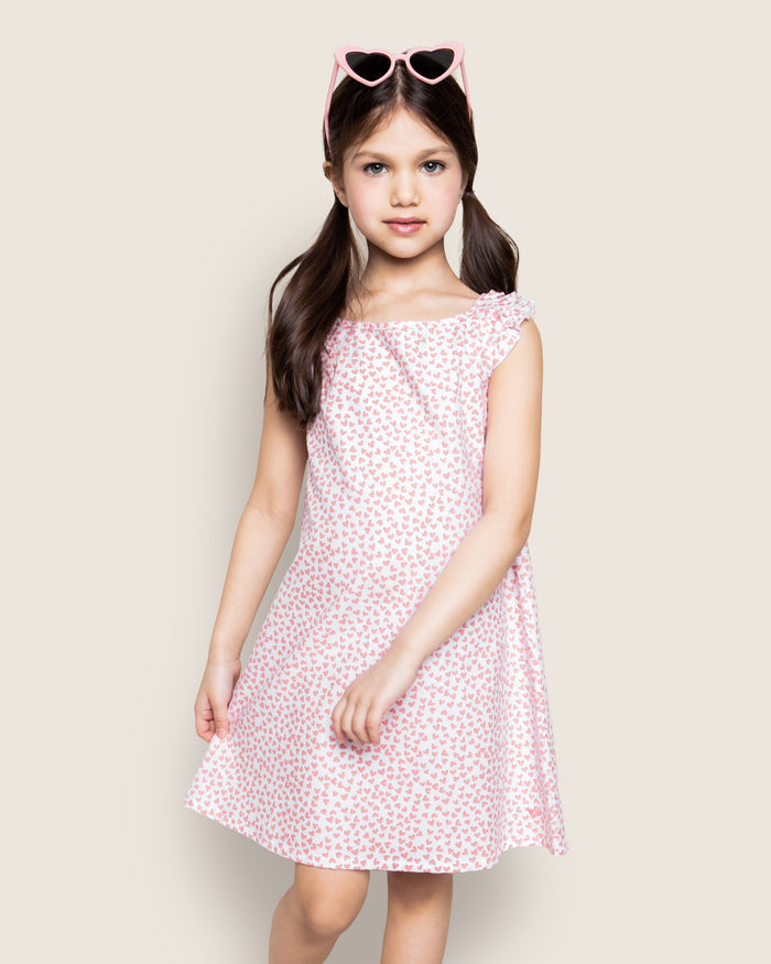 A young girl with long brown pigtails stands wearing the sleeveless Girls Twill Amelie Nightgown in Sweethearts by Petite Plume. She sports heart-shaped sunglasses on her head, set against a plain, light background.