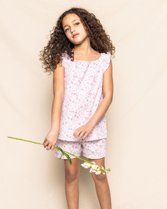A young girl with curly hair wears the Petite Plume Girls Twill Amelie Short Set in Dorset Floral, crafted from soft cotton. She holds a single stem of white flowers and stands gracefully against a neutral background.