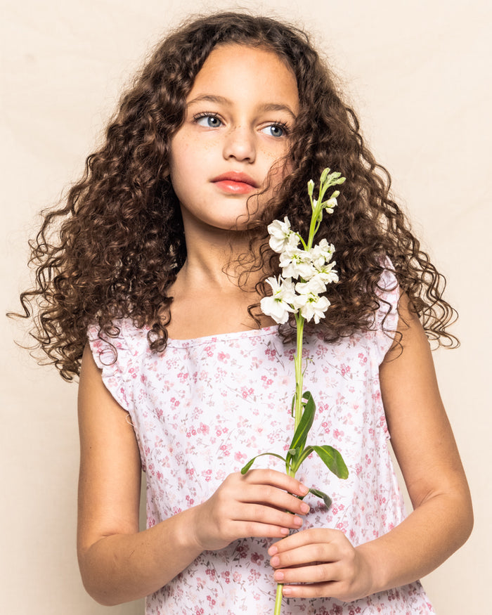 A young girl with curly brown hair holds white flower buds. She wears the Girls Twill Amelie Short Set in Dorset Floral by Petite Plume and gazes thoughtfully to the side against a neutral background.