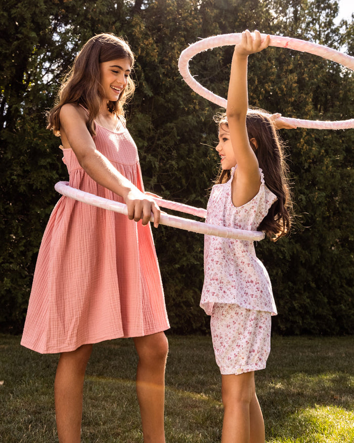 Two young girls are playing with hula hoops on a grassy area. A girl in a pink dress helps another wearing the Girls Twill Amelie Short Set in Dorset Floral by Petite Plume hold a hula hoop over her head. Both smile, surrounded by lush greenery and flower buds.