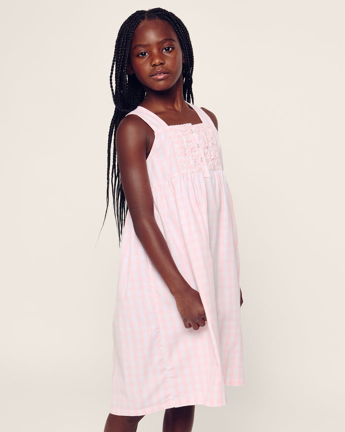 Four individuals in light-colored sleepwear pose on a stone wall in a sunny, wooded area. Two sit relaxed, and two stand below. One wears the Petite Plume Girls Twill Charlotte Nightgown in Pink Gingham, highlighting the natural and comforting setting of their attire.