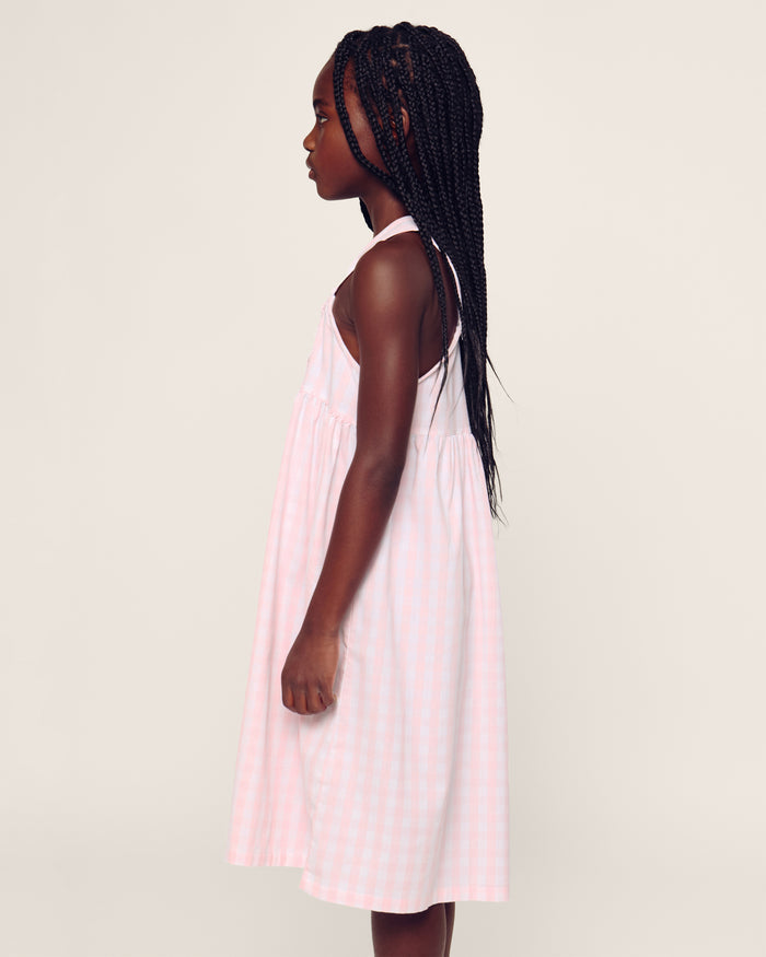 Four individuals in light-colored sleepwear pose on a stone wall in a sunny, wooded area. Two sit relaxed, and two stand below. One wears the Petite Plume Girls Twill Charlotte Nightgown in Pink Gingham, highlighting the natural and comforting setting of their attire.