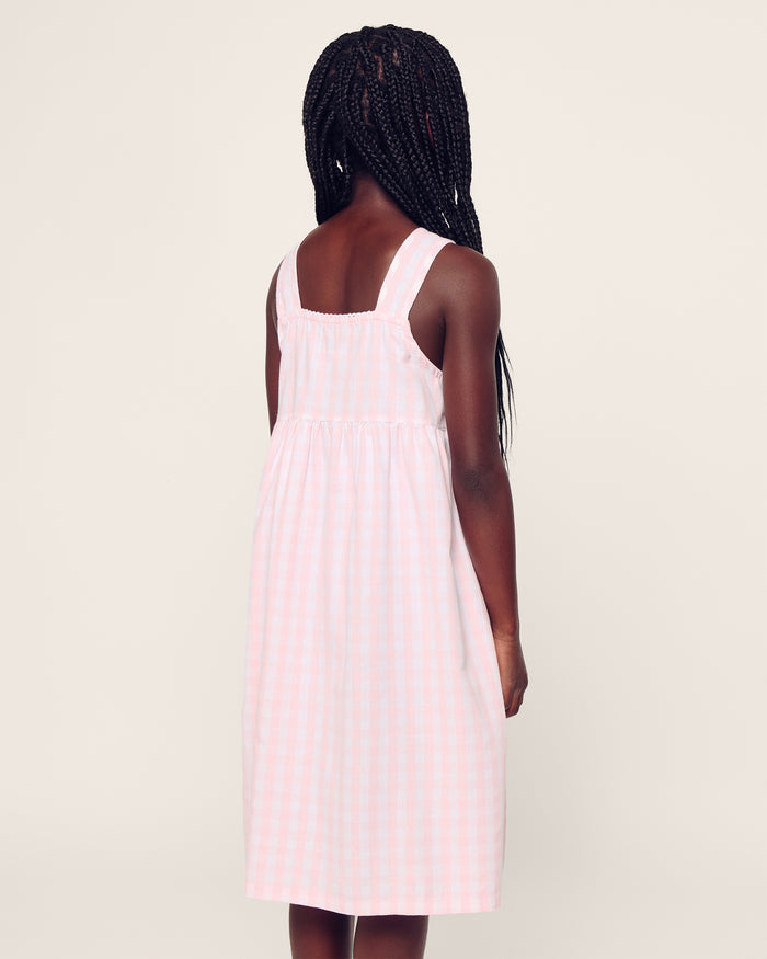 Four individuals in light-colored sleepwear pose on a stone wall in a sunny, wooded area. Two sit relaxed, and two stand below. One wears the Petite Plume Girls Twill Charlotte Nightgown in Pink Gingham, highlighting the natural and comforting setting of their attire.