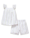 A young girl with curly hair smiles barefoot, wearing the Girl's Twill Clara Short Set in Cottage Stripe— a white sleeveless nightgown and matching shorts—against a plain, light background.