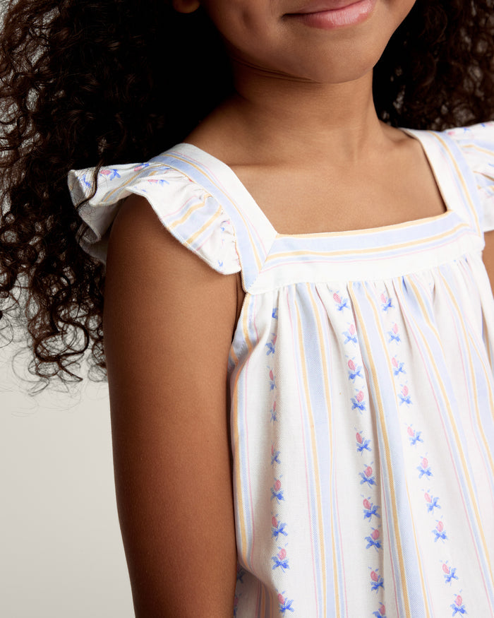 A young girl with curly hair smiles barefoot, wearing the Girl's Twill Clara Short Set in Cottage Stripe— a white sleeveless nightgown and matching shorts—against a plain, light background.