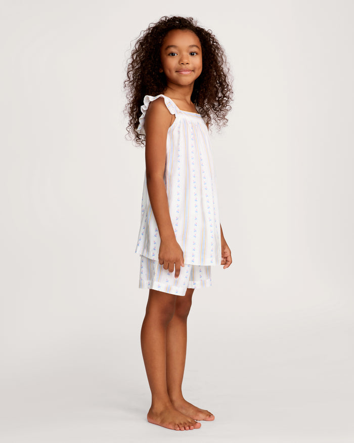 A young girl with curly hair smiles barefoot, wearing the Girl's Twill Clara Short Set in Cottage Stripe— a white sleeveless nightgown and matching shorts—against a plain, light background.