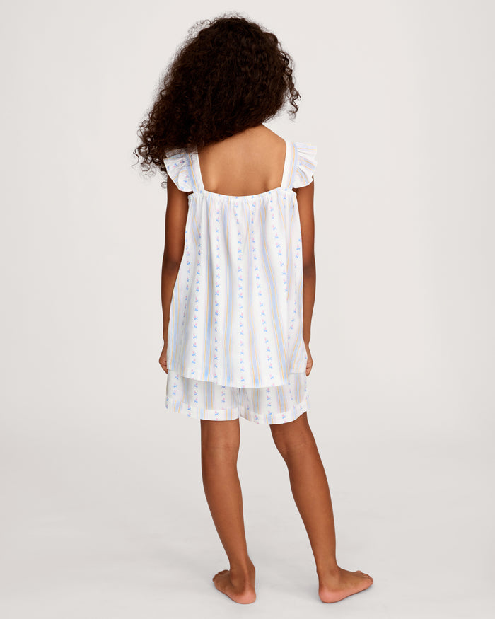 A young girl with curly hair smiles barefoot, wearing the Girl's Twill Clara Short Set in Cottage Stripe— a white sleeveless nightgown and matching shorts—against a plain, light background.