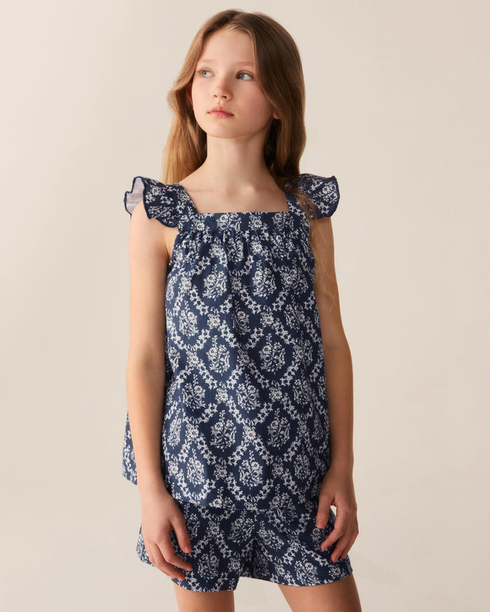 A young girl with long brown hair wears the Girl’s Twill Clara Short Set in Innkeeper Trellis—a blue top and matching shorts featuring a white floral pattern. She stands against a plain light background, looking slightly to her right with a neutral expression.