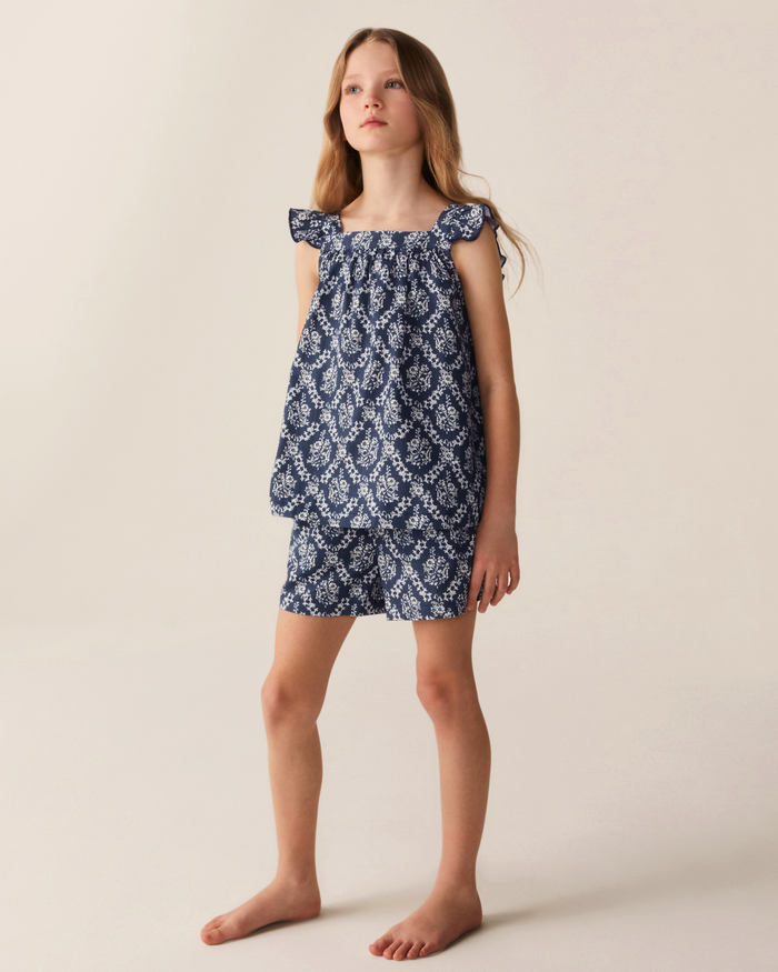 A young girl stands barefoot against a plain light background, wearing the Girl’s Twill Clara Short Set in Innkeeper Trellis—a matching blue and white patterned sleeveless top with ruffled straps and coordinating shorts.