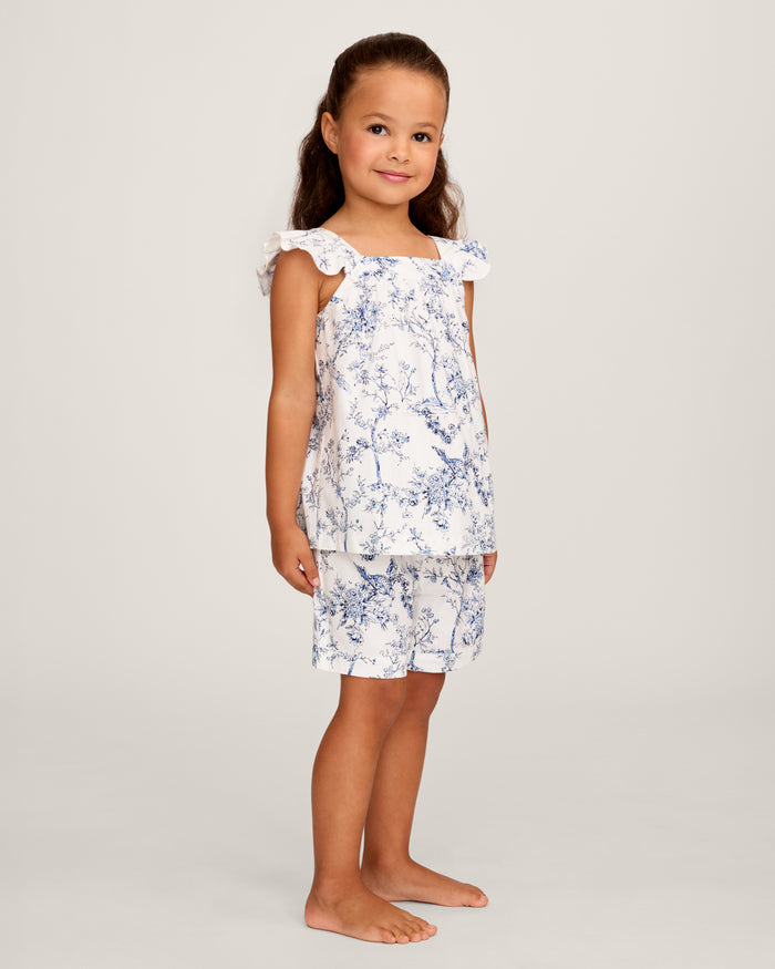 A young girl with long brown hair stands barefoot, smiling in the Girl's Twill Clara Short Set in Timeless Toile—a white top and matching blue floral shorts—posing against a plain light background.