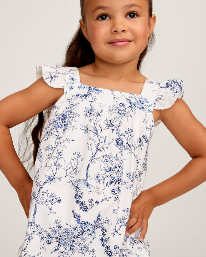 A young girl with long brown hair stands barefoot, smiling in the Girl's Twill Clara Short Set in Timeless Toile—a white top and matching blue floral shorts—posing against a plain light background.