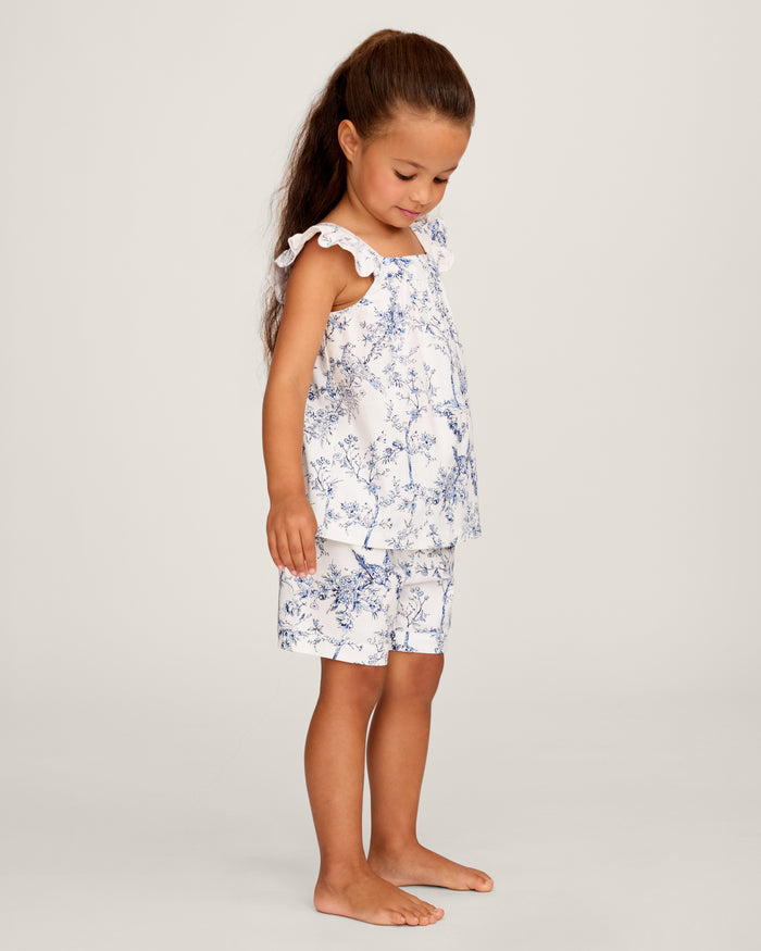 A young girl with long brown hair stands barefoot, smiling in the Girl's Twill Clara Short Set in Timeless Toile—a white top and matching blue floral shorts—posing against a plain light background.