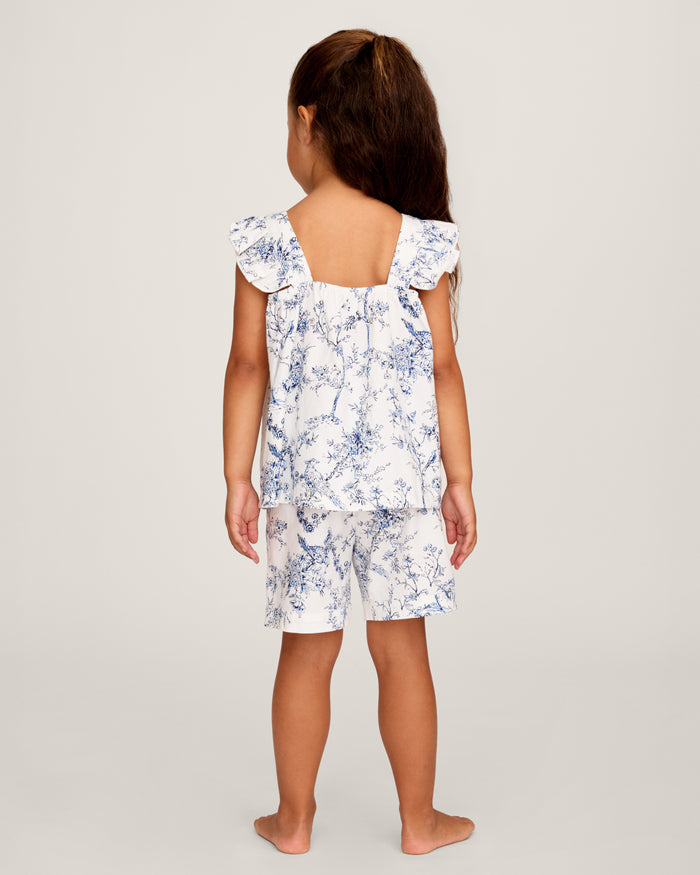 A young girl with long brown hair stands barefoot, smiling in the Girl's Twill Clara Short Set in Timeless Toile—a white top and matching blue floral shorts—posing against a plain light background.