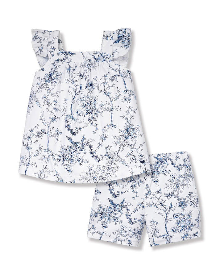 A young girl with long brown hair stands barefoot, smiling in the Girl's Twill Clara Short Set in Timeless Toile—a white top and matching blue floral shorts—posing against a plain light background.