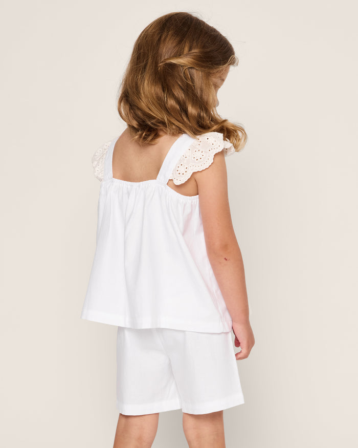 A young girl with brown hair stands facing away, wearing Petite Plumes Girls Twill Clara Short Set in white. The finest quality cotton features lace details on the sleeveless top, perfectly matched with shorts against a plain background.