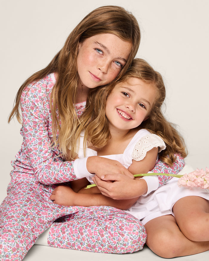 Two smiling young girls embrace. The older one, with long brown hair, wears Petite Plumes Girls Twill Clara Short Set in White. The younger girl holds pink flowers and wears a white dress. Both sit against a light background.