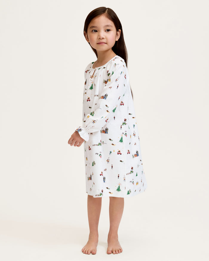 The Girl's Twill Delphine Nightgown in Holiday Town is a white, long-sleeved children's nightgown with colorful festive prints and features ruffled cuffs and a gathered neckline.