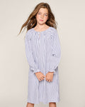 A young girl with long brown hair wearing Petite Plumes Girls Twill Delphine Nightgown in Navy French Ticking, crafted from flame-retardant fabric, stands against a plain background with a relaxed expression, facing forward.