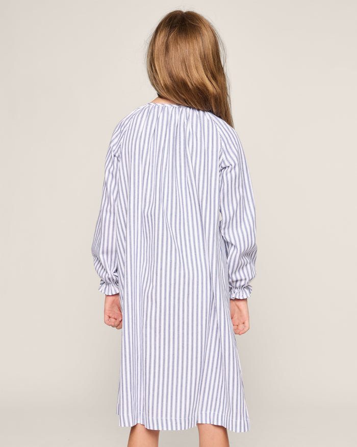A young girl with long hair facing away wears Petite Plumes Girls Twill Delphine Nightgown in Navy French Ticking, made from flame-retardant fabric. The sleepwear features vertical blue and white stripes against a plain, light-colored background.