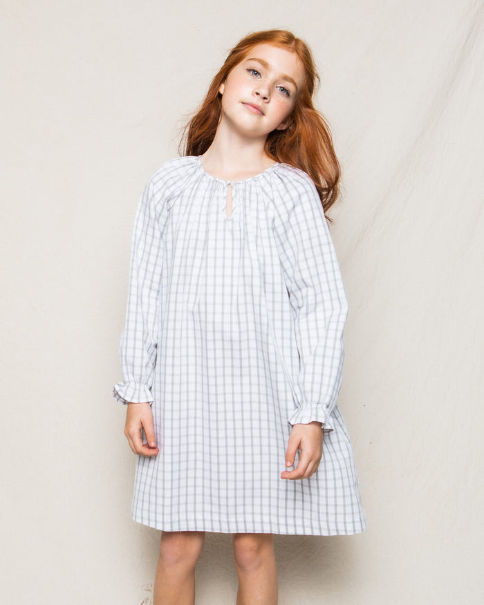 A young girl with long red hair stands against a plain backdrop in Petite Plumes Girls Twill Delphine Nightgown in Regent Tattersall, reminiscent of luxurious sleepwear. She tilts her head slightly, gazing forward with serene confidence.