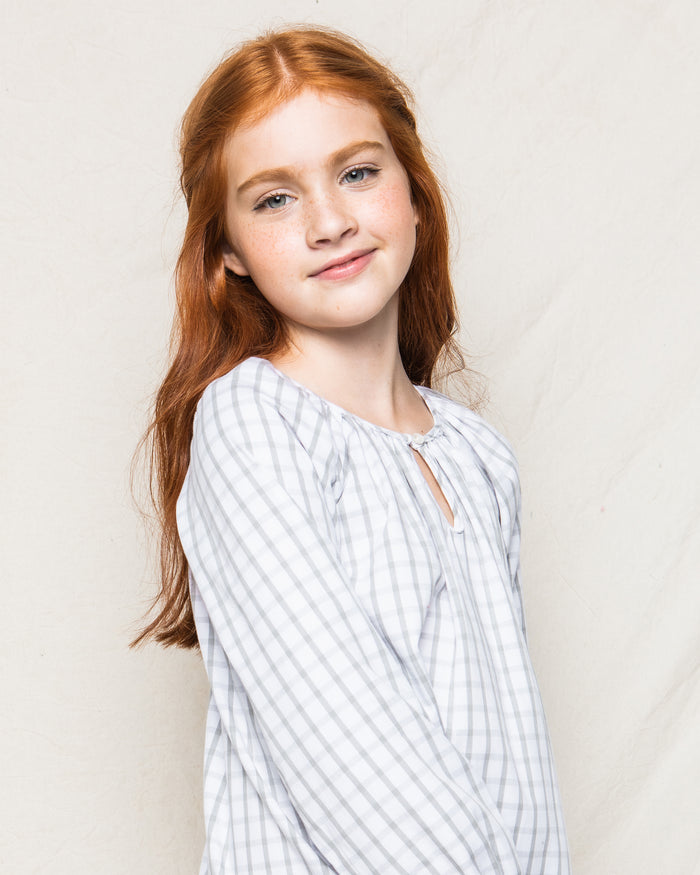 A young girl with long red hair and freckles softly smiles at the camera, wearing Petite Plumes Girls Twill Delphine Nightgown in Regent Tattersall, set against a plain cream-colored background.