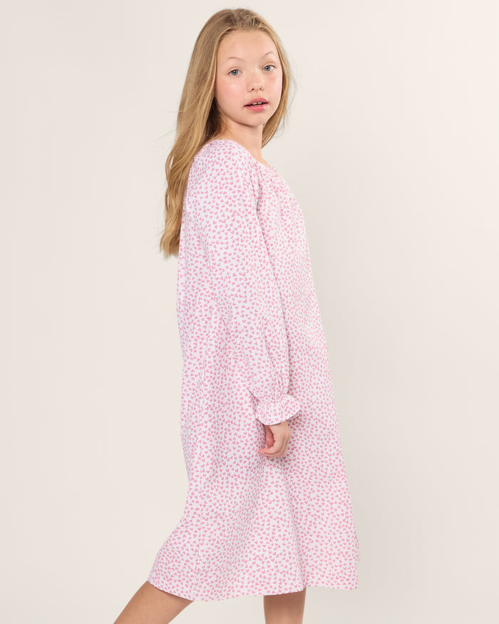 A young girl with long blonde hair stands sideways in a Petite Plume Girls Twill Delphine Nightgown in Sweethearts, looking towards the camera with a neutral expression against a plain, light-colored background.