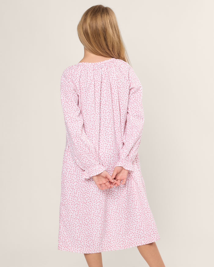 A girl with long blonde hair stands with her back to the camera, wearing a Petite Plume Girls Twill Delphine Nightgown in Sweethearts. The gowns long sleeves drape gracefully as she clasps her hands behind her back against an off-white background.