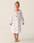 A young girl stands barefoot in the Girl’s Twill Delphine Nightgown in Trick or Treat, a white, long-sleeve nightgown adorned with a charming print. She gazes slightly to the side and smiles softly against a plain, light background.