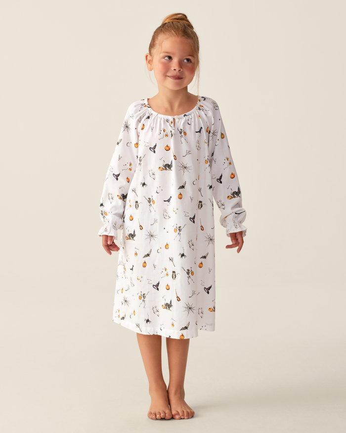 A young girl stands barefoot in the Girl’s Twill Delphine Nightgown in Trick or Treat, a white, long-sleeve nightgown adorned with a charming print. She gazes slightly to the side and smiles softly against a plain, light background.