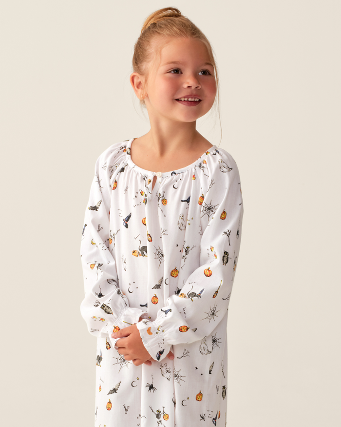 A young girl with light brown hair styled in a bun is smiling and looking to the side against a plain background, wearing the Girl’s Twill Delphine Nightgown in Trick or Treat, which features playful Halloween-themed prints.