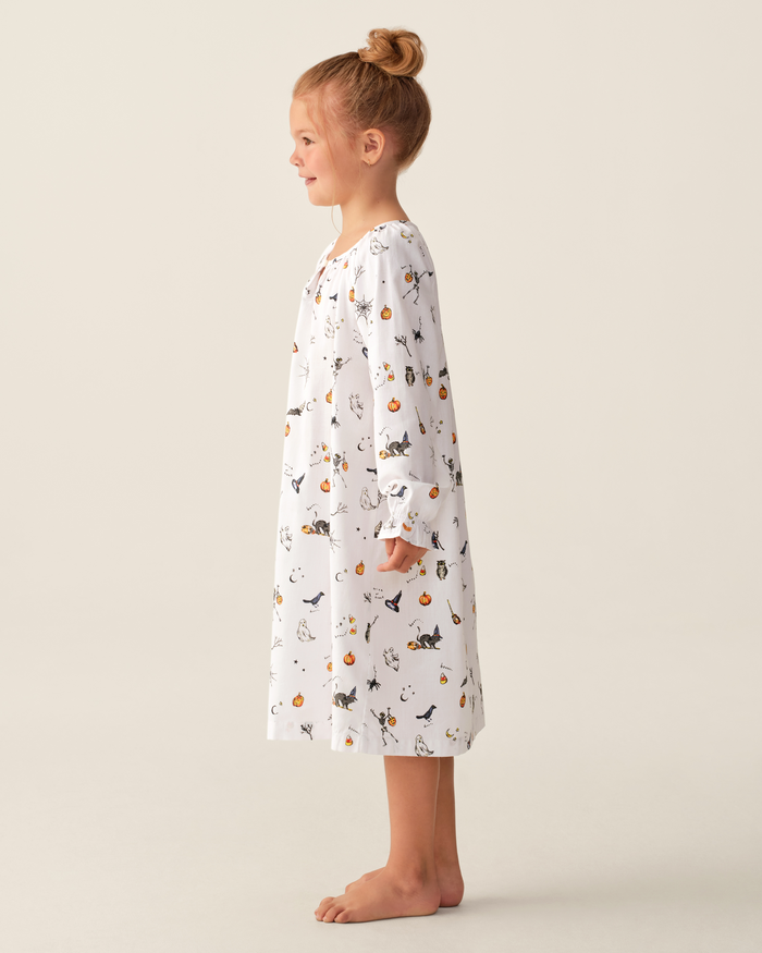 A young girl stands barefoot in profile, wearing the Girl’s Twill Delphine Nightgown in Trick or Treat—a long-sleeve white nightgown adorned with playful, colorful animal and pumpkin prints. Her hair is styled in a neat bun, set against a plain light beige background.