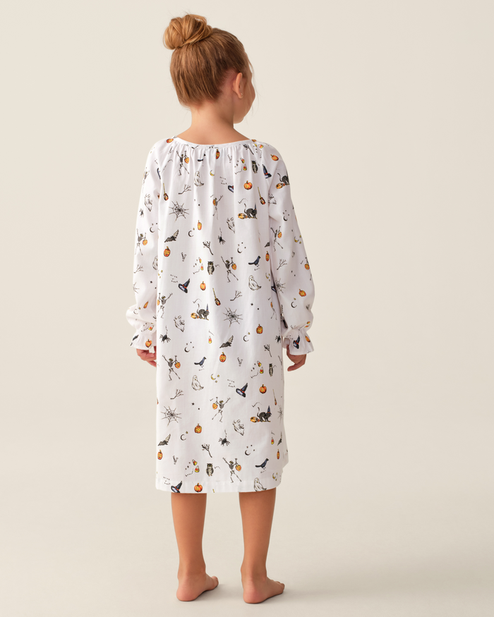 A young girl with light brown hair in a bun stands barefoot and faces away, wearing the Girl’s Twill Delphine Nightgown in Trick or Treat—a long-sleeved white nightgown adorned with a Halloween-themed print of pumpkins, cats, and bats—against a plain light background.