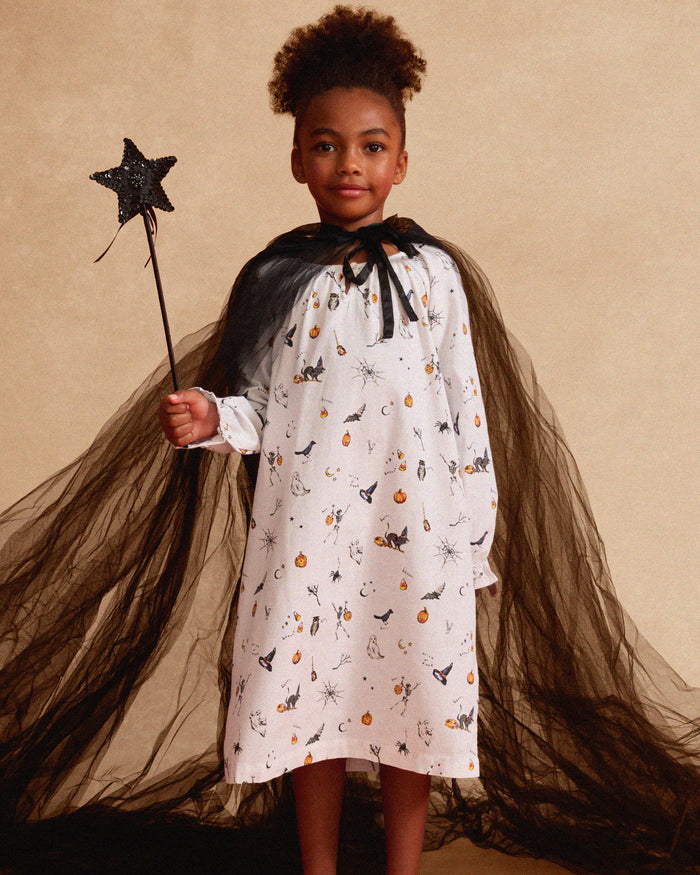 The Petite Plume Girls Twill Delphine Nightgown in Trick or Treat showcases Halloween-themed prints like pumpkins, ghosts, and bats. This white long-sleeve gown features gathered sleeves and a button-fastened neckline, ideal for the festive season.