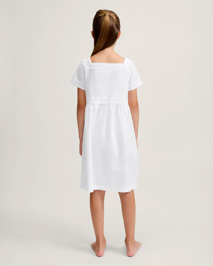 A young girl stands barefoot against a light background, wearing the Girl's Twill Juliette Nightgown in White—a knee-length piece with short sleeves, lace trim, and pintuck details. She gazes gently to the side.