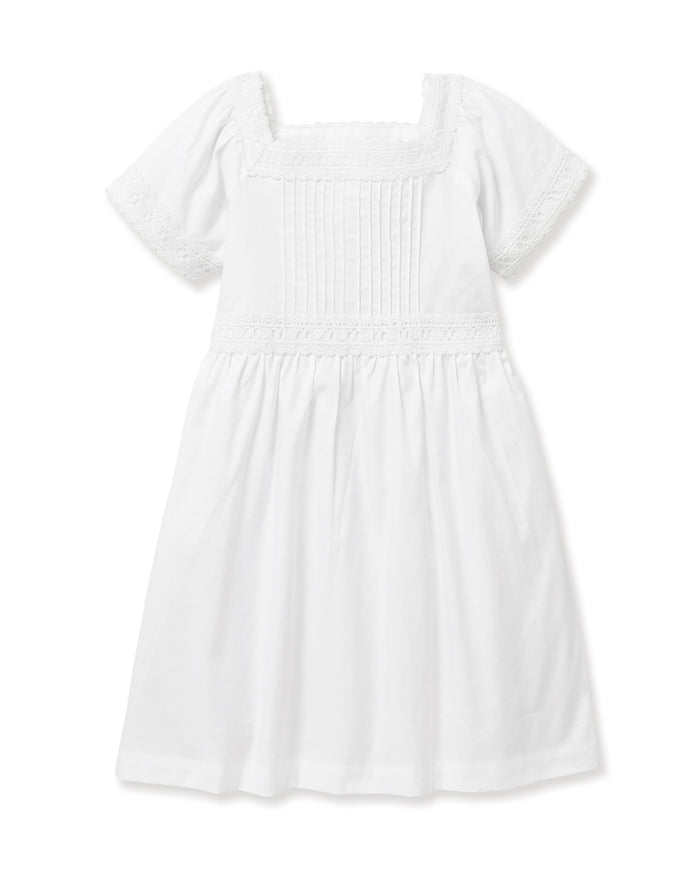 A young girl stands barefoot against a light background, wearing the Girl's Twill Juliette Nightgown in White—a knee-length piece with short sleeves, lace trim, and pintuck details. She gazes gently to the side.