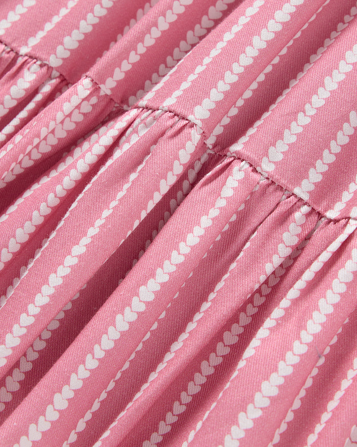 The Girls Twill Lilly Nightgown in Love Lines by Petite Plume features pink and white striped fabric with small heart patterns. The gathered yarn-dyed cotton creates ruffled folds, perfect for luxurious sleepwear.