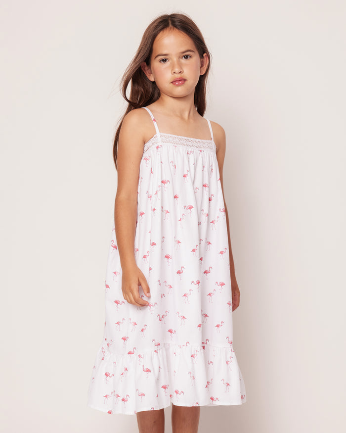 A young girl with long brown hair wears Petite Plumes Girls Twill Lily Nightgown in Flamingos, featuring thin straps and a ruffled hem. She stands against a plain off-white background with a neutral expression, the small pink flamingo pattern adding a tropical twist.