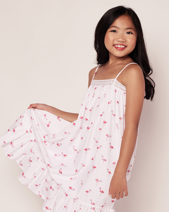 A young girl, with long dark hair, smiles while holding out the skirt of her Girls Twill Lily Nightgown in Flamingos by Petite Plume against a light background.