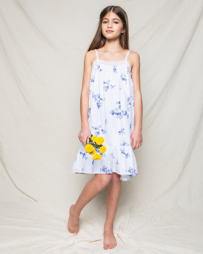 A young girl in Petite Plumes Indigo Floral Twill Lily Nightgown stands barefoot against a light background, holding a bunch of yellow flowers.