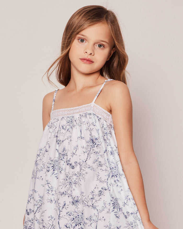 A girl with long brown hair wears the Girls Twill Lily Nightgown in Timeless Toile by Petite Plume. Standing against a plain backdrop, she exudes botanical elegance as she looks at the camera. The white gown features blue floral patterns, thin straps, and lace detailing, offering luxurious sleepwear style.