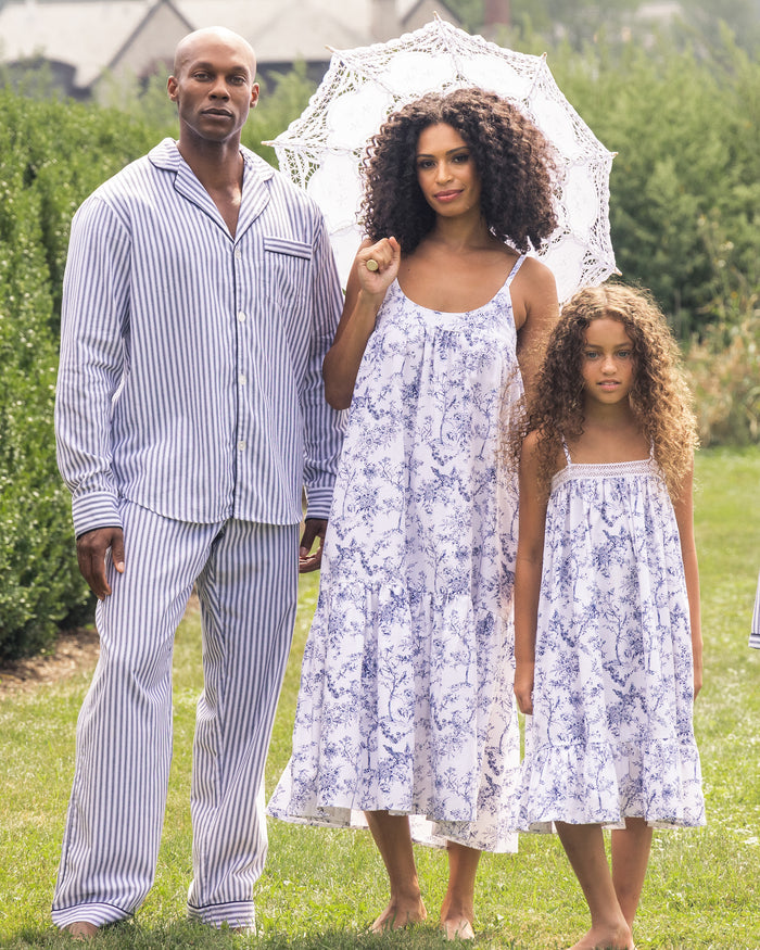 A family is outdoors on a sunny day, radiating botanical elegance. The father wears striped pajamas, while the mother holds a white parasol and dons a floral dress that matches her childs Petite Plume Girls Twill Lily Nightgown in Timeless Toile. Vibrant greenery enhances the scene.
