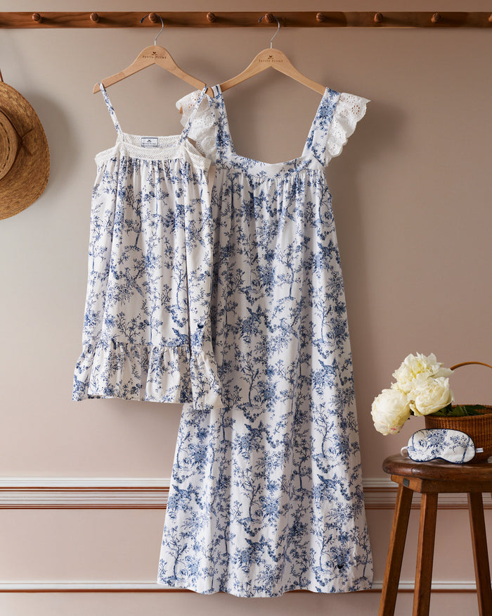 Two luxury Girls Twill Lily Nightgowns in Timeless Toile by Petite Plume hang on wooden hangers against a beige wall. A straw hat is on the left, and white flowers sit on a stool to the right, with a matching sleep mask beside them. Crafted from flame retardant cotton for safety and style.