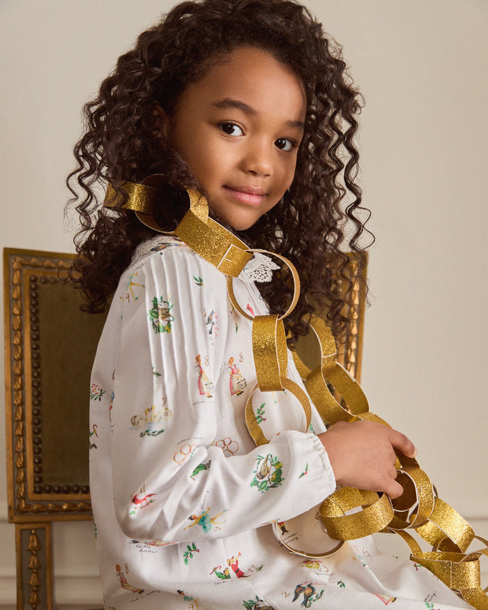 The Girl's Twill Lola Nightgown in 12 Days of Christmas is a white long-sleeve children’s dress with a Peter Pan collar and a playful pattern of colorful people, animals, and plants scattered across the fabric.