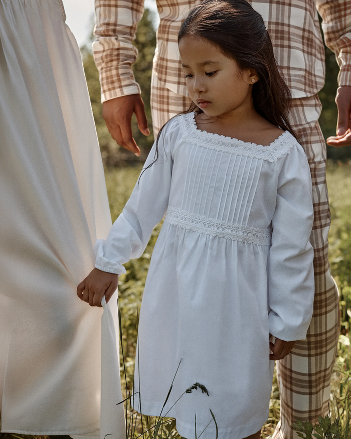 A young girl wearing the Girl’s Twill Long Sleeve Juliette Nightgown in White stands outdoors, holding the hand of an adult beside her. Two adults dressed in white and plaid are partially visible in the background, surrounded by greenery.