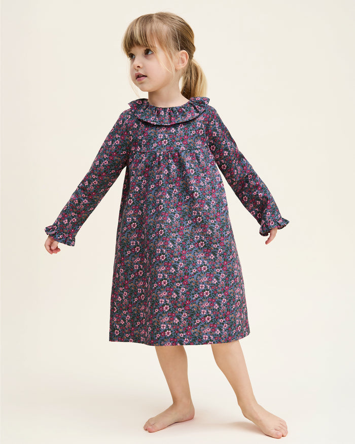 The Girl's Twill Scarlett Nightgown in Fleurs de Saison is a long-sleeved, knee-length dress with a ruffled collar and cuffs, featuring pink and white flowers with green leaves on a dark background.