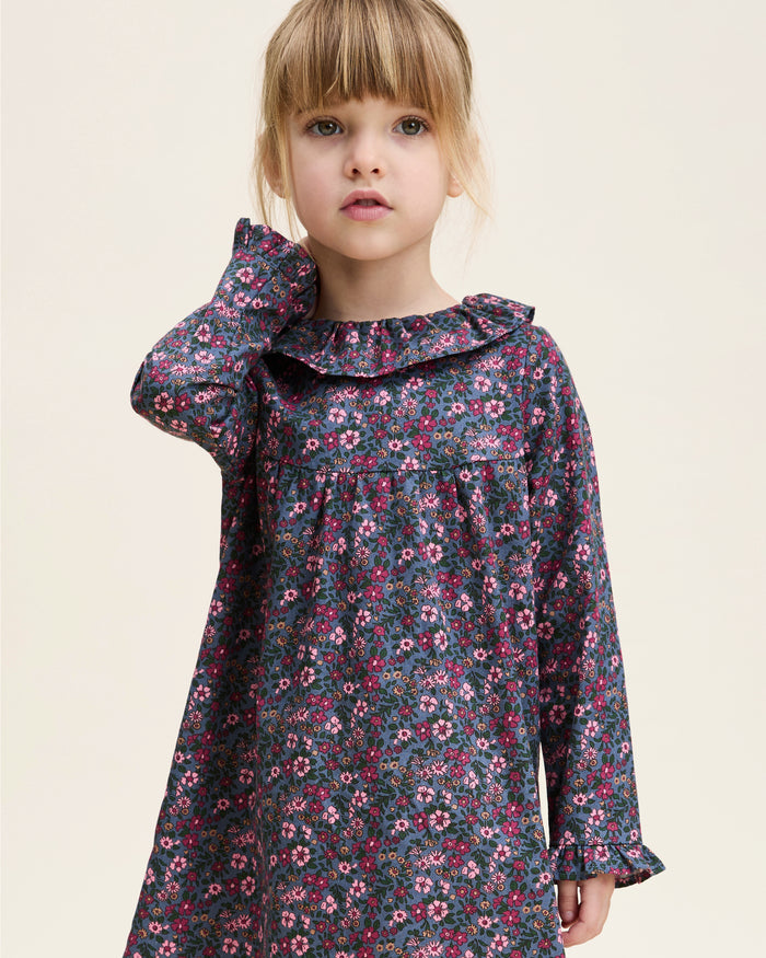 The Girl's Twill Scarlett Nightgown in Fleurs de Saison is a long-sleeved, knee-length dress with a ruffled collar and cuffs, featuring pink and white flowers with green leaves on a dark background.