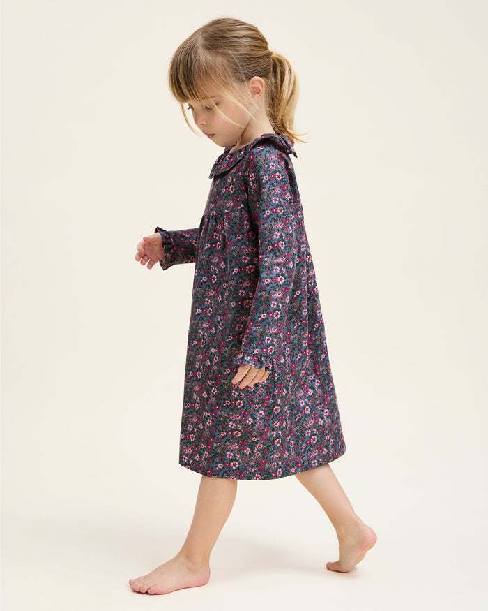 The Girl's Twill Scarlett Nightgown in Fleurs de Saison is a long-sleeved, knee-length dress with a ruffled collar and cuffs, featuring pink and white flowers with green leaves on a dark background.