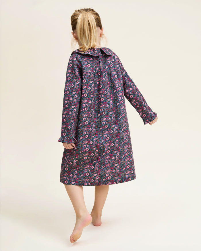 The Girl's Twill Scarlett Nightgown in Fleurs de Saison is a long-sleeved, knee-length dress with a ruffled collar and cuffs, featuring pink and white flowers with green leaves on a dark background.