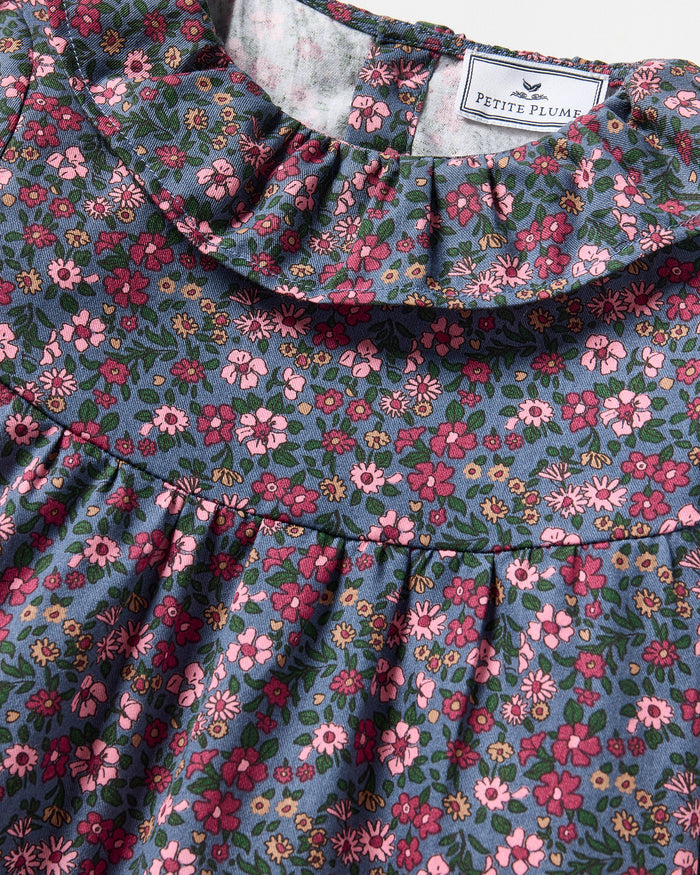 The Girl's Twill Scarlett Nightgown in Fleurs de Saison is a long-sleeved, knee-length dress with a ruffled collar and cuffs, featuring pink and white flowers with green leaves on a dark background.