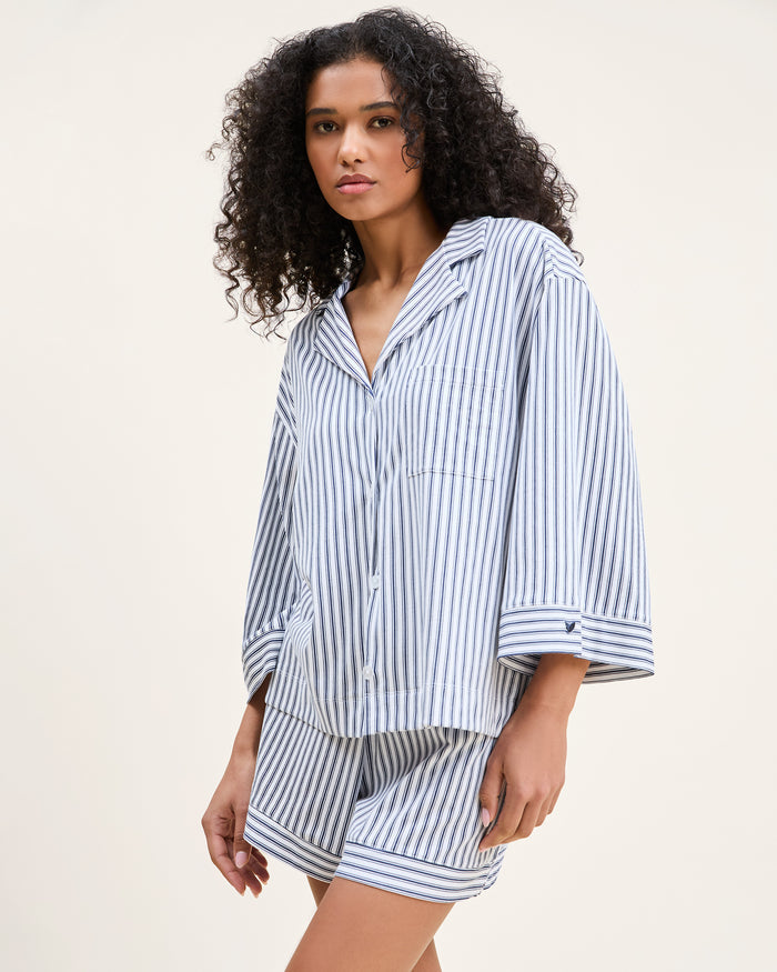 The Olivia Pima Short Set in Navy French Ticking features a matching set of blue and white vertical striped pajamas with a long-sleeve, button-up shirt with chest pocket and drawstring shorts.
